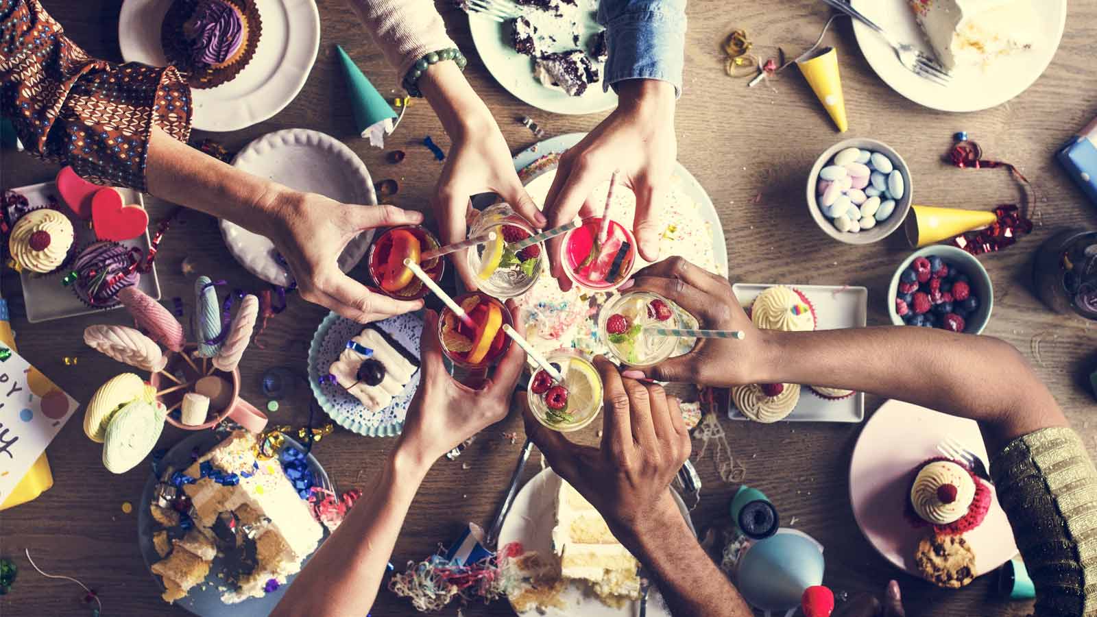 A birds-eye view of friends clinking cocktails over an afternoon tea cake banquet